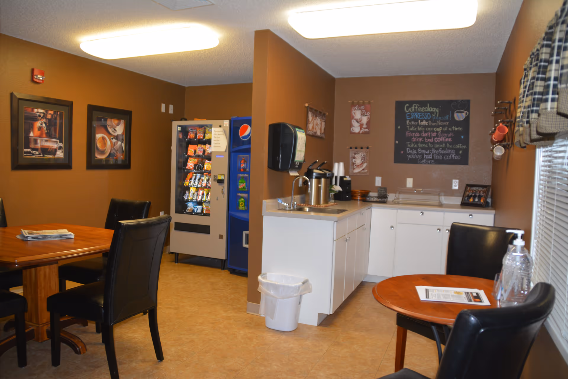 A cozy common area with brown walls featuring a snack vending machine and a beverage cooler in the corner. There are two wooden tables with black leather chairs around them. One table has newspapers on it, and the other has a hand sanitizer bottle and a paper. A small kitchenette area with a sink, coffee dispensers, cups, and a chalkboard with coffee-related sayings is visible. The room is warmly lit with ceiling lights and has framed coffee-themed artwork on the walls.