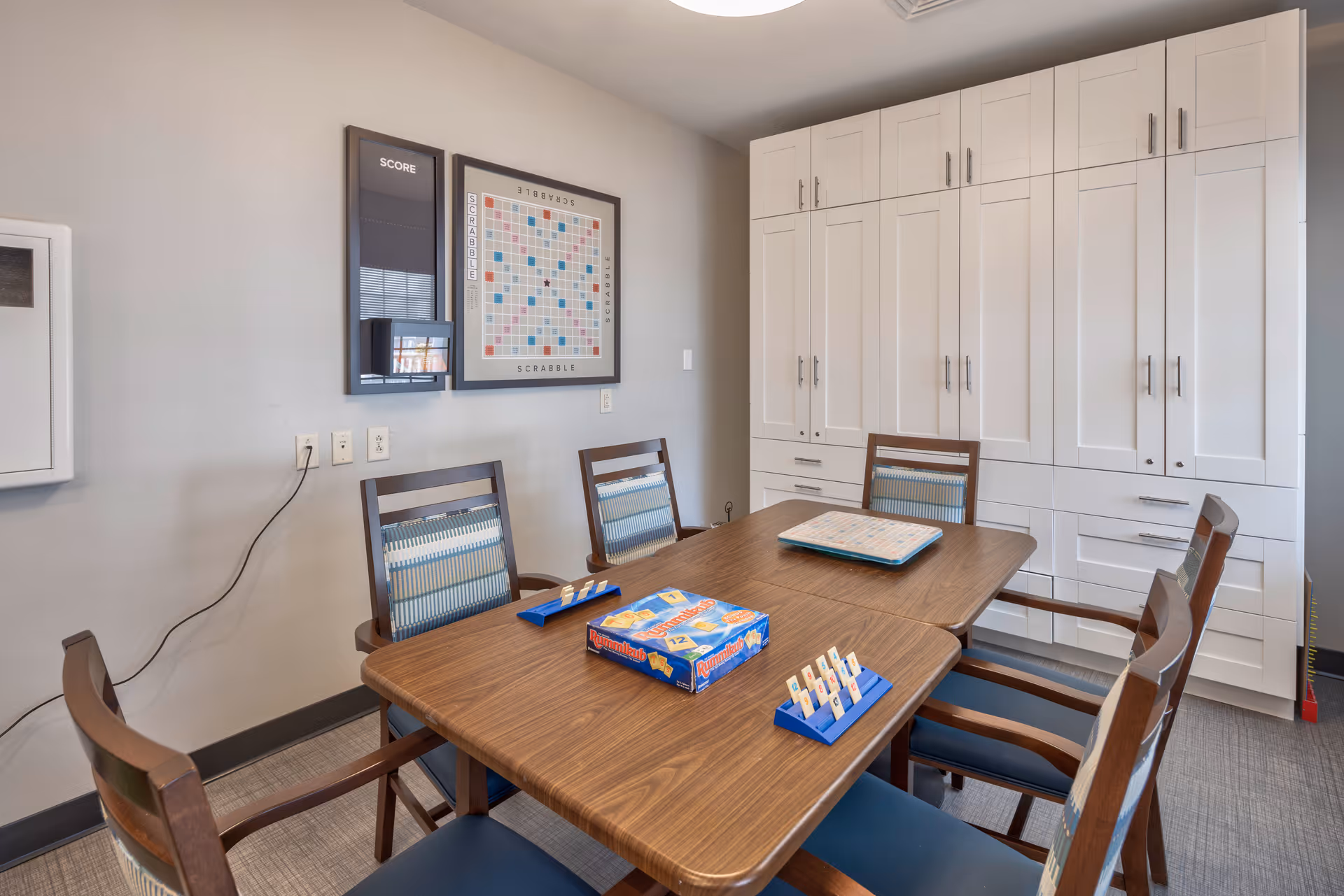 Interior activity room with a wooden table surrounded by chairs, Scrabble tiles and racks on the table, and tall white storage cabinets against the wall.