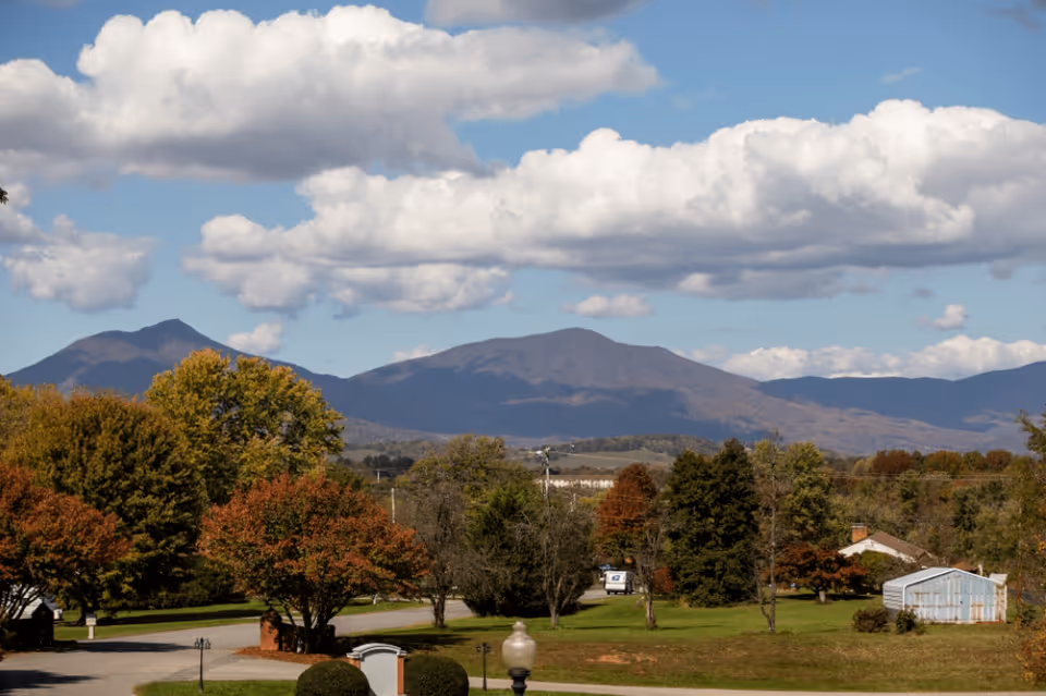 A scenic outdoor view featuring a variety of trees with autumn foliage, a grassy area, a few buildings including a small shed, and mountains in the background under a partly cloudy blue sky.