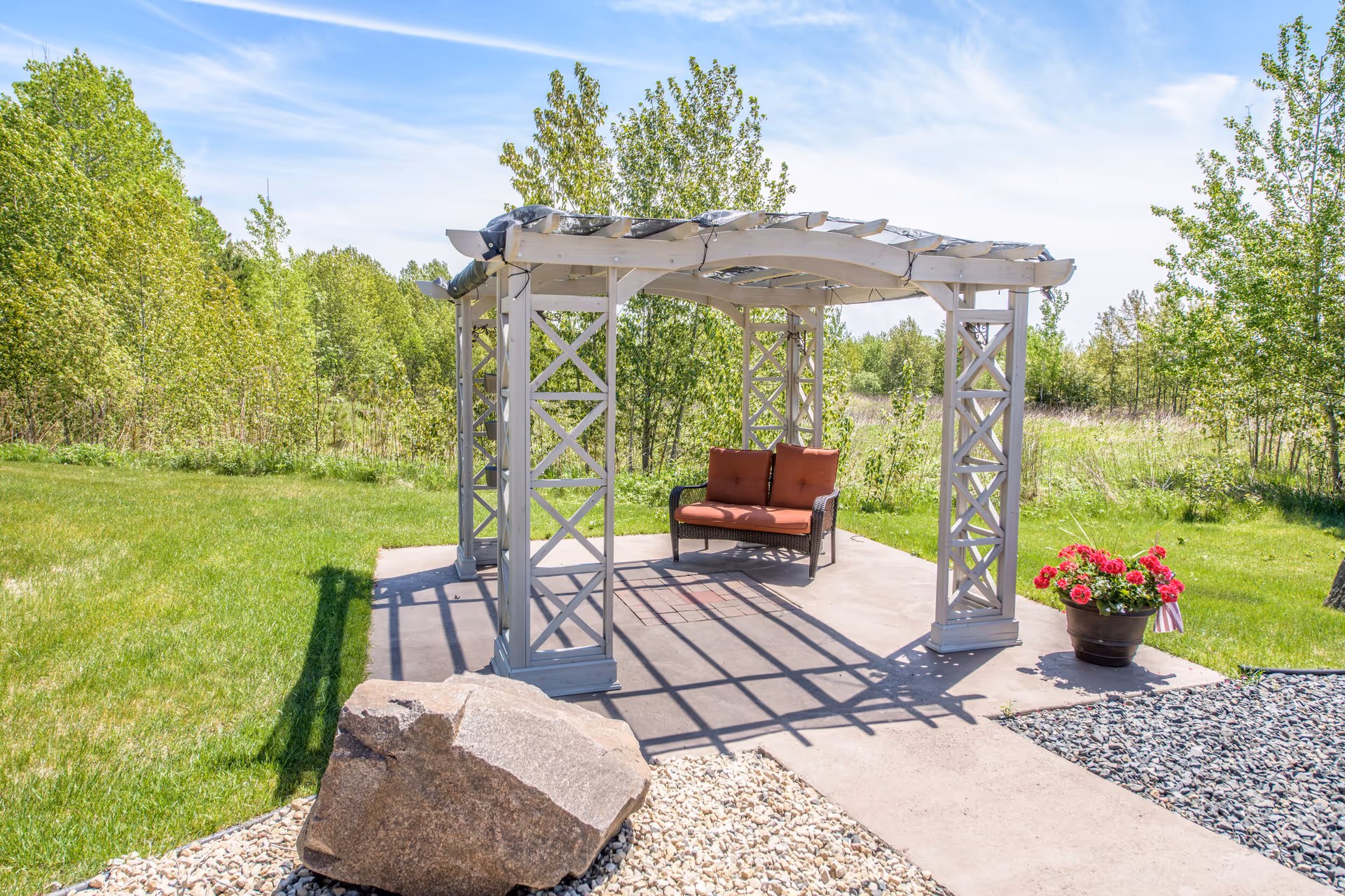 A white wooden pergola with a cushioned bench on a concrete patio surrounded by lawn, trees, and a potted flower.