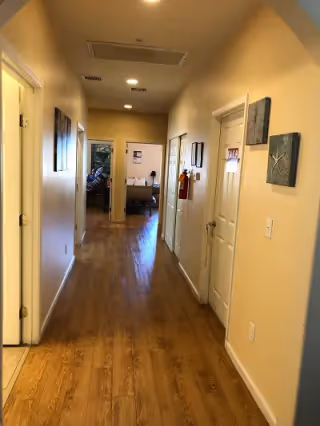 A hallway with wooden flooring and beige walls, featuring several closed white doors on both sides. At the end of the hallway, a room is visible with a bed and some furniture. The hallway is lit by ceiling lights and decorated with framed artwork on the walls.
