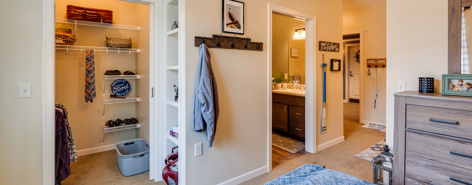 Interior view of a senior living facility bedroom area showing an open walk-in closet with shelves and hanging clothes, a coat rack with a jacket, a bathroom with a sink and mirror, and part of a dresser with framed photos and decorative items.