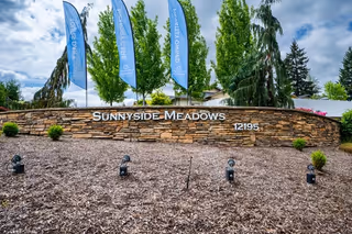 Stone wall with the sign 'Sunnyside Meadows' and the number '12195' in front of tall green trees and blue flags under a partly cloudy sky.