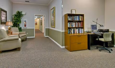 A quiet interior hallway area in a senior living facility with beige walls and carpeted floor. On the left side, there are two beige armchairs with a lamp and a potted plant. On the right side, there is a wooden bookshelf filled with books and a desk with a computer and office chair. The hallway extends into the distance with framed artwork on the walls.