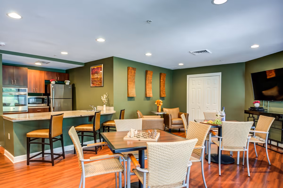 A cozy common area with a small kitchen featuring stainless steel appliances and wooden cabinets. There are bar stools at the kitchen counter, a table with a chessboard and chairs, and another table with chairs and a bottle of wine with glasses. The walls are painted green with decorative wall hangings and a painting. There is a TV mounted on the wall and comfortable armchairs near a set of white double doors.