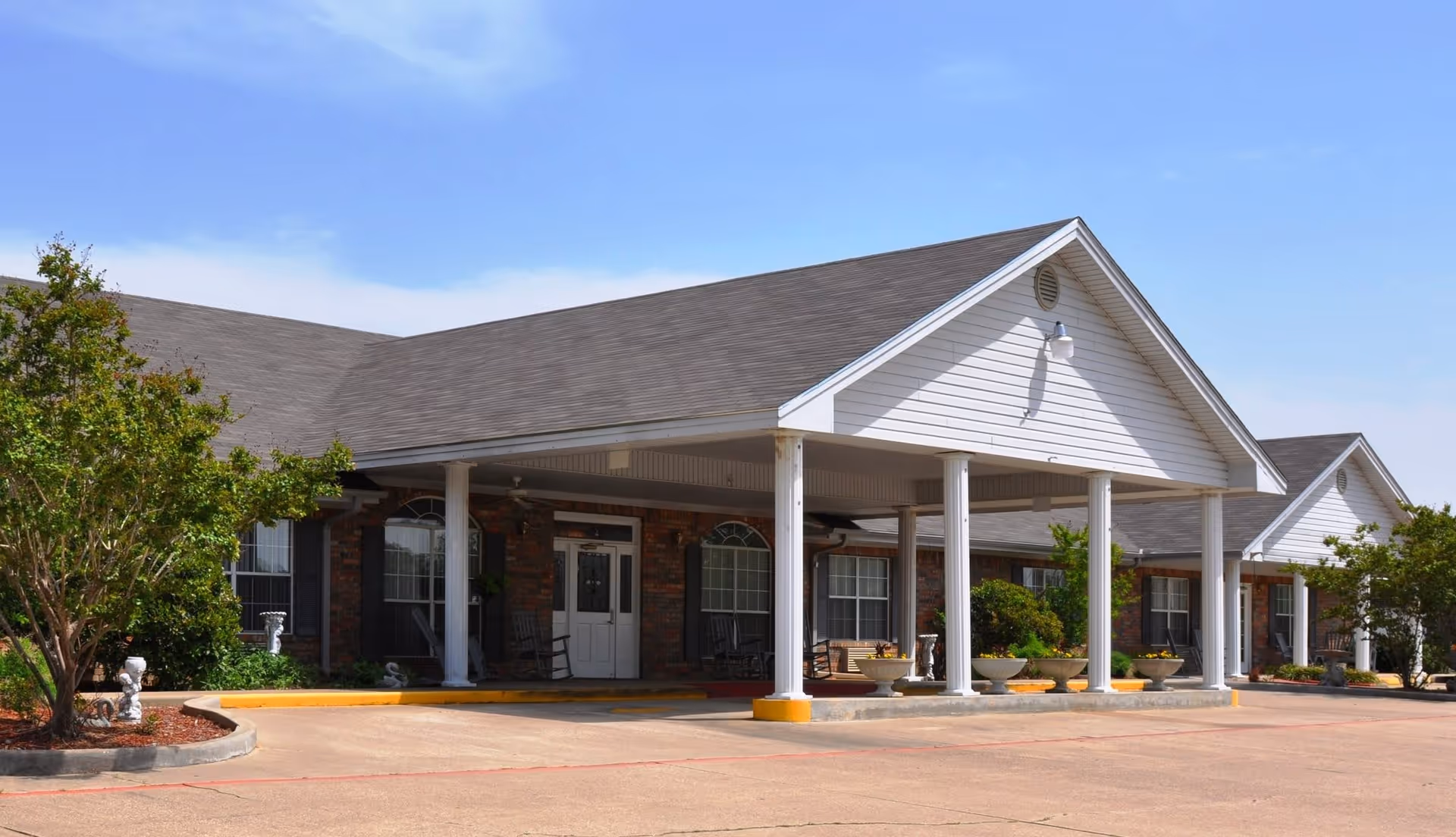 Exterior view of The Meadows senior living facility showing a single-story brick building with white columns supporting a covered entrance. There are several windows with black shutters, rocking chairs on the porch, and landscaped greenery around the building under a clear blue sky.