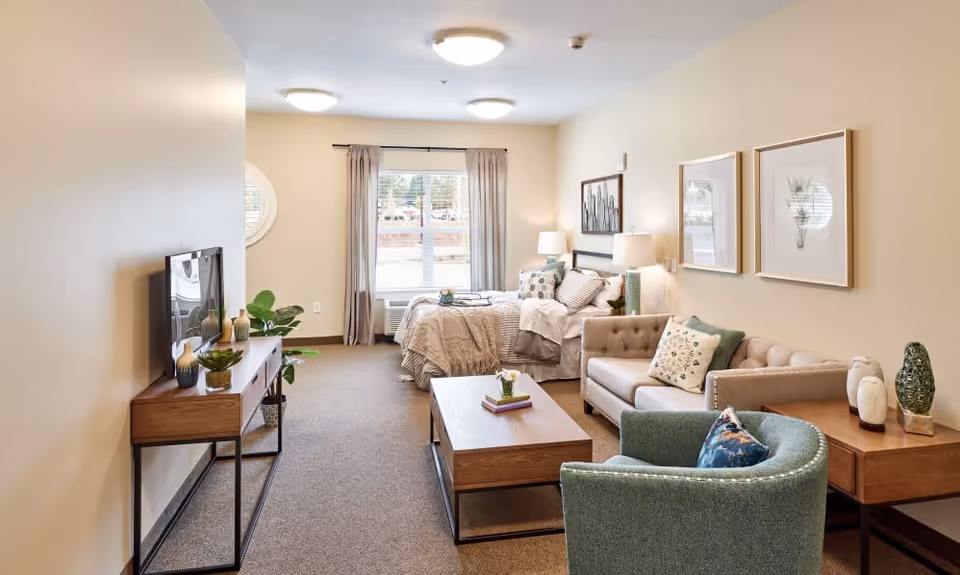 A cozy and well-lit room with a bed near a window with curtains, a beige loveseat with decorative pillows, a green armchair, a wooden coffee table with books and a small plant, a wooden console table with a TV and decorative items, and framed artwork on the walls.