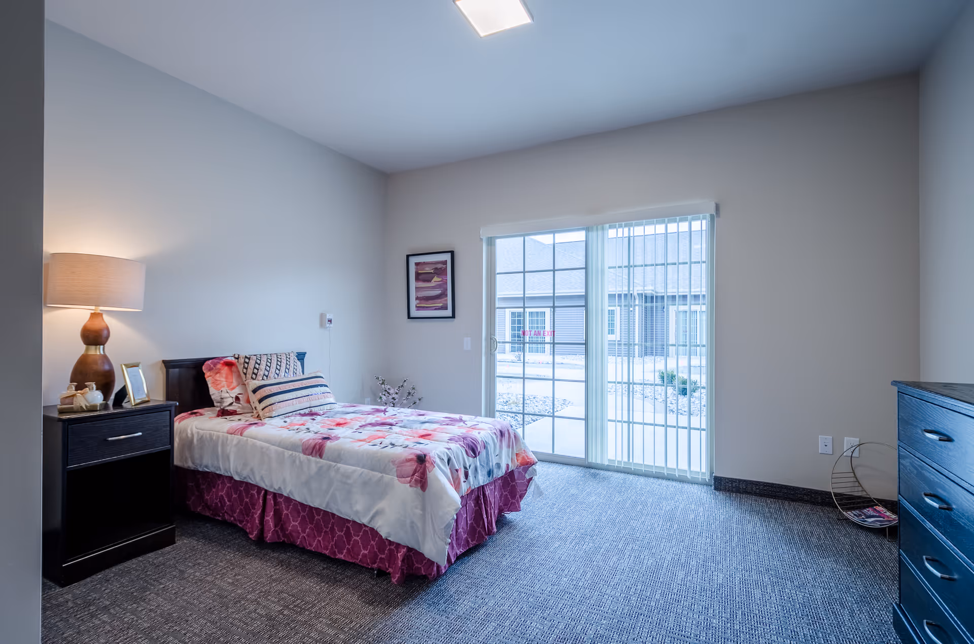 A bedroom with a single bed covered in floral bedding and multiple pillows. There is a nightstand with a lamp and decorative items on the left side of the bed. A framed artwork hangs on the wall above the bed's headboard. To the right, there is a large sliding glass door with vertical blinds that opens to an outdoor area. A dark wooden dresser is visible on the right side of the room, and the floor is carpeted.