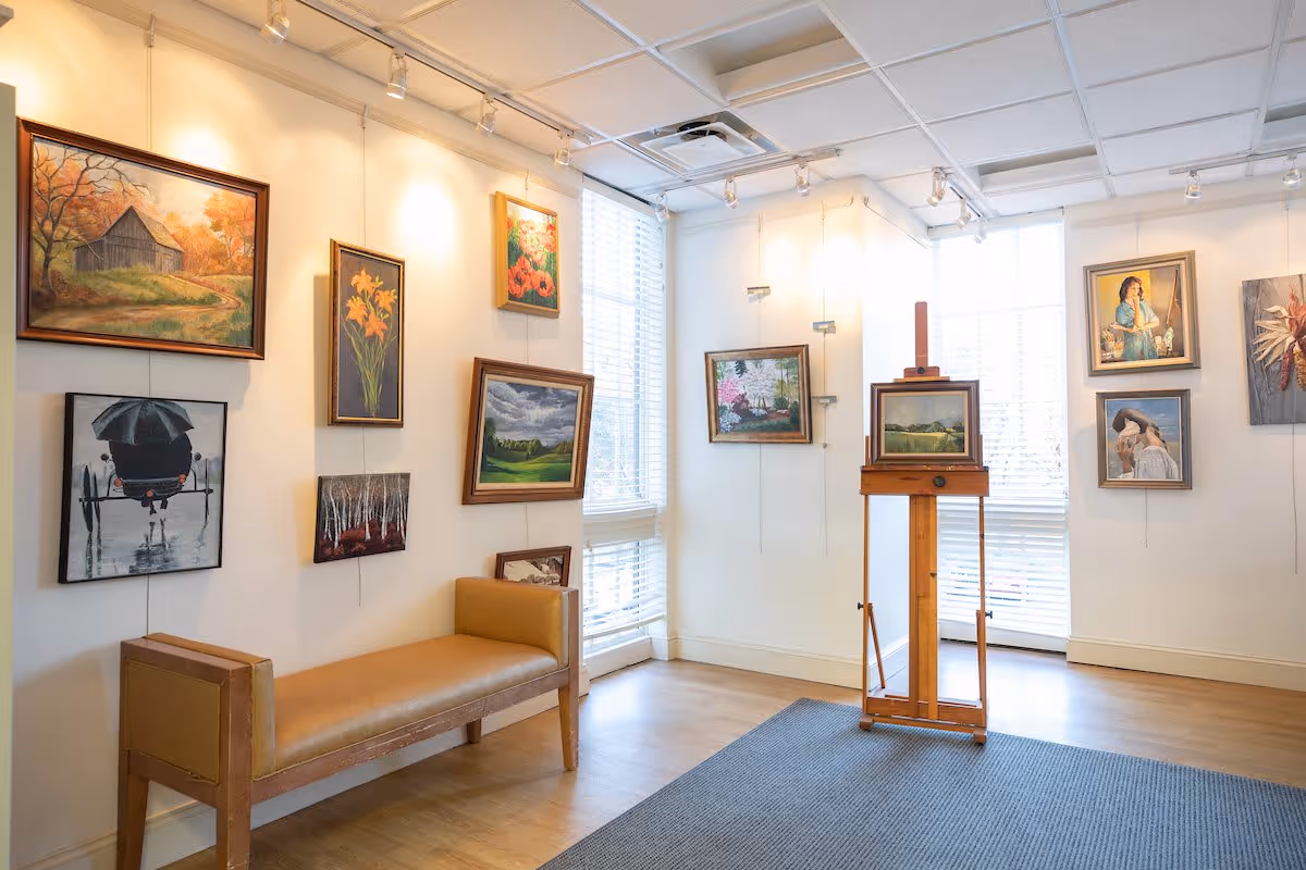 A bright art gallery room with several framed paintings hanging on white walls. There is a wooden bench with a tan cushion along one wall and a wooden easel displaying a landscape painting near large windows with white blinds. The floor is light wood with a gray rug in the center.