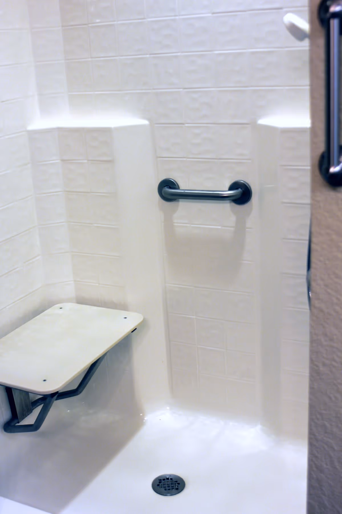 A white tiled shower area with a foldable shower seat mounted on the wall and a horizontal grab bar for support.
