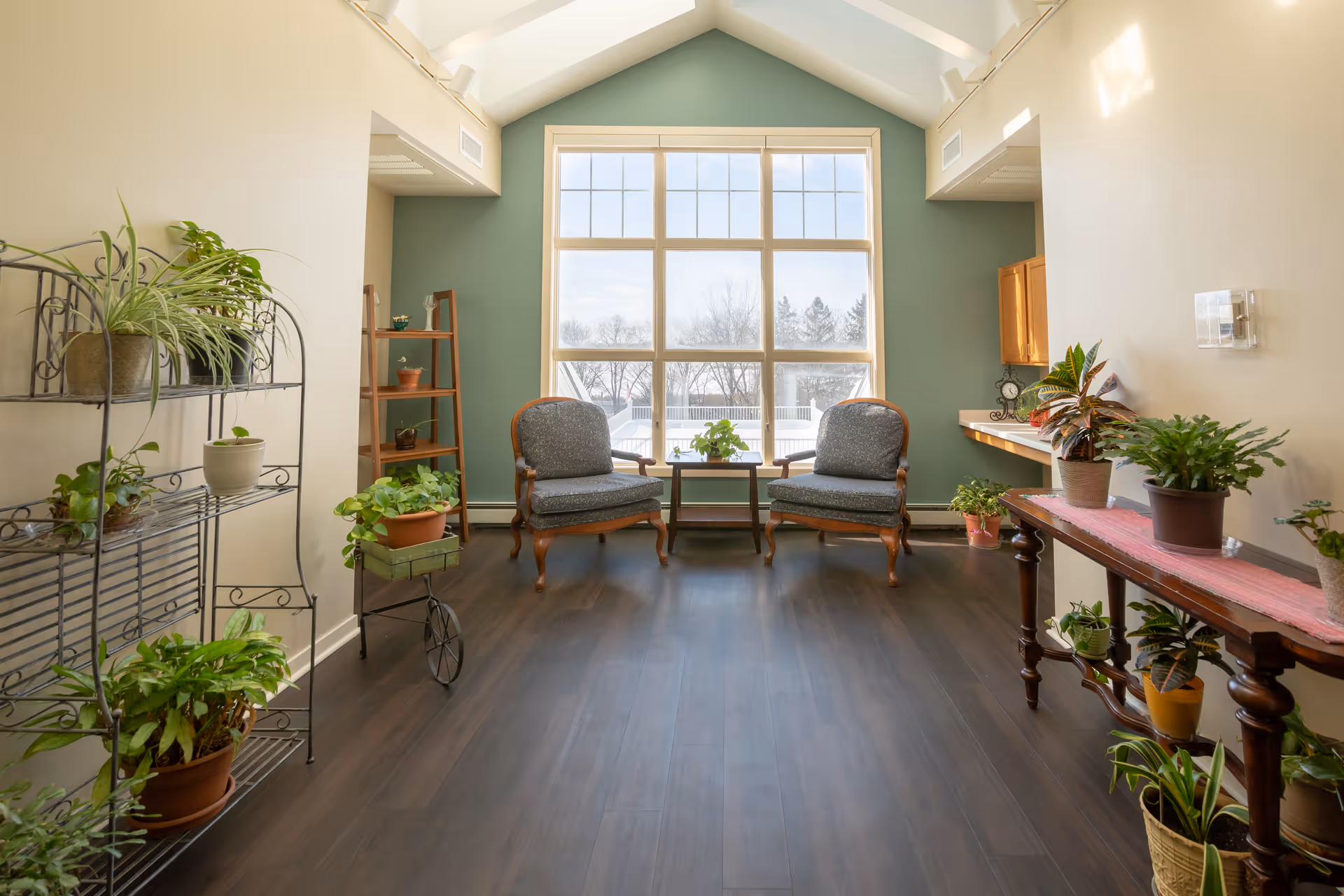 Sunlit common sitting area with two armchairs facing a large window, hardwood floors, and numerous potted plants.