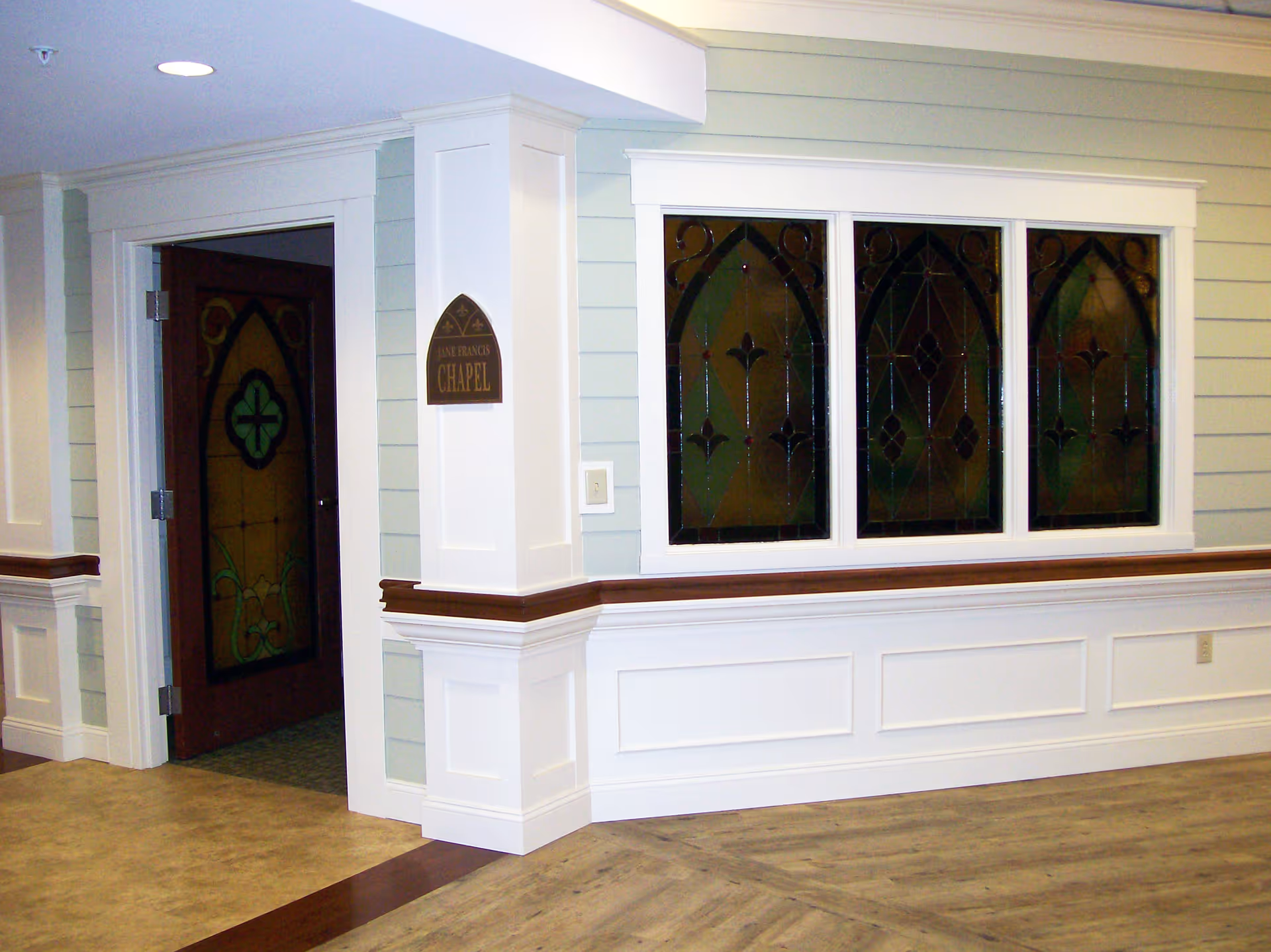 Interior hallway corner with light green walls and white wainscoting, featuring a wooden door with stained glass leading to a chapel, and three stained glass windows on the adjacent wall.