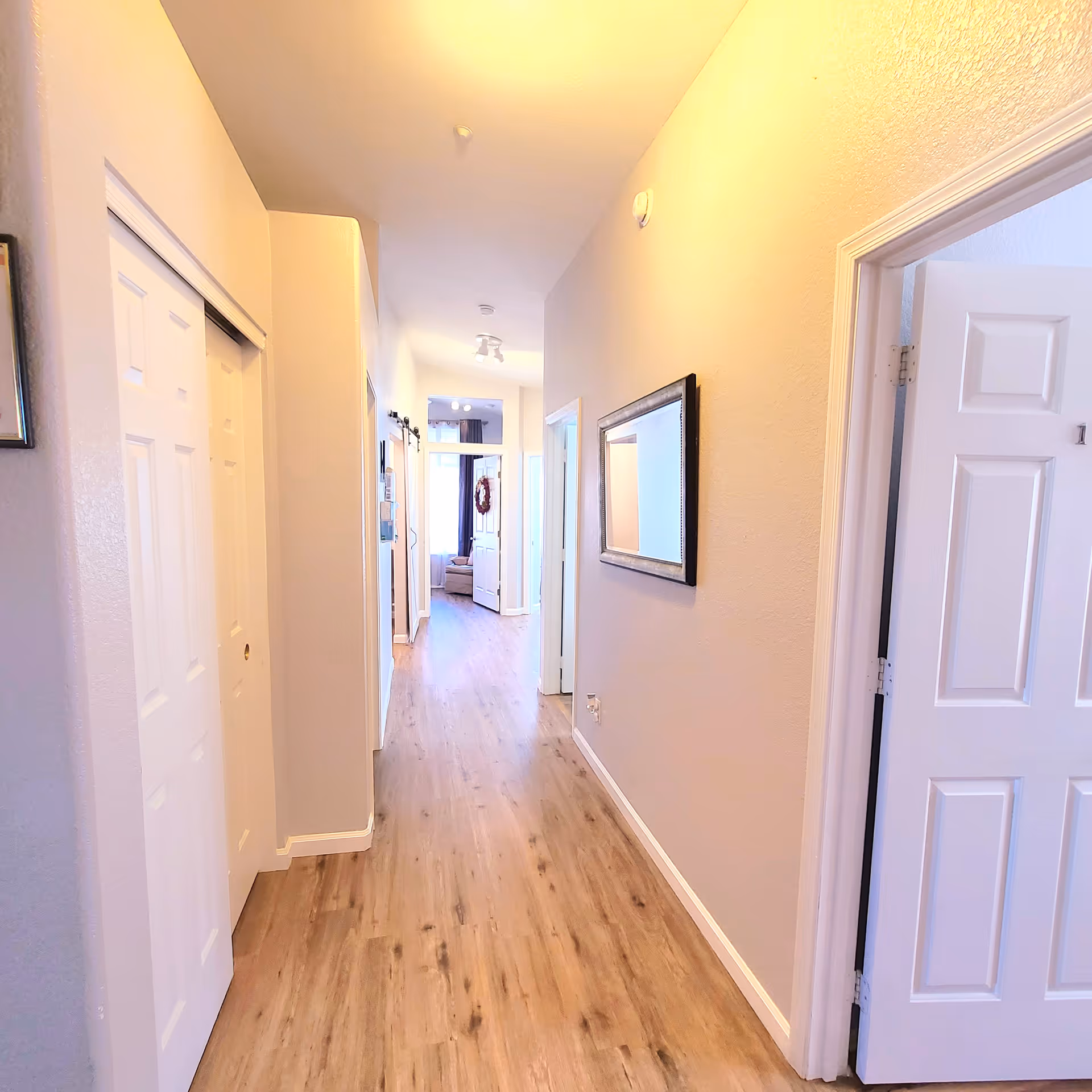 A bright hallway with light-colored walls and wood flooring. There are white doors on both sides of the hallway, a framed mirror on the right wall, and a glimpse of a living area with a window and curtains at the far end.