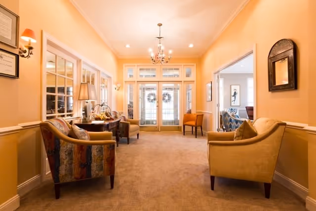 A warmly lit sitting area with beige walls and carpet, featuring four upholstered armchairs arranged around a wooden console table with a lamp and decorative items. The space has a chandelier hanging from the ceiling and large glass doors with wreaths leading outside. There are framed pictures and a mirror on the walls.