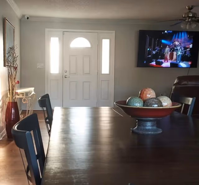 Dark wood dining table with chairs and a decorative bowl in the foreground, a front entry door and a wall-mounted TV visible in the background.