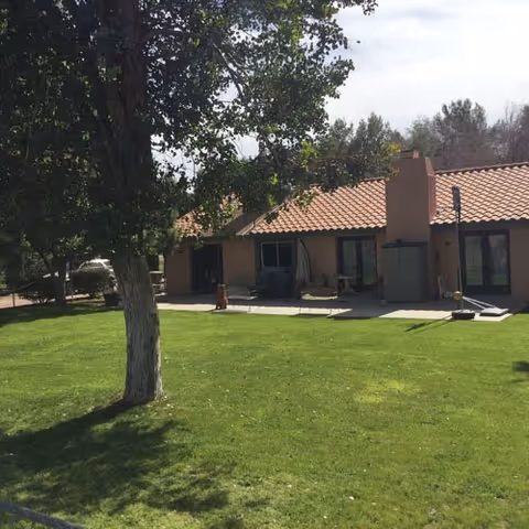 A single-story building with a tiled roof and multiple glass doors opening to a patio area. The patio has outdoor furniture and a barbecue grill. In the foreground, there is a well-maintained green lawn with a large tree providing shade.