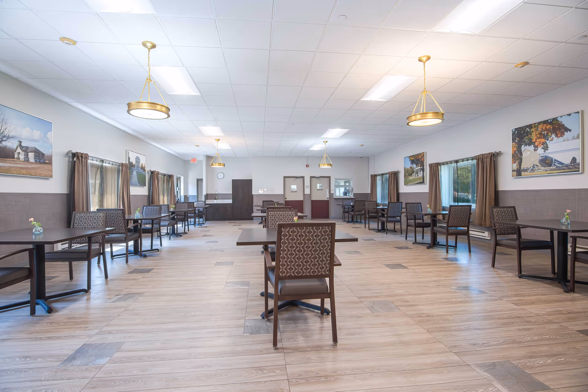 A spacious dining room with multiple tables and chairs arranged neatly. The room has wooden flooring, large windows with brown curtains, and several framed pictures on the walls. Ceiling lights with gold fixtures hang evenly spaced across the ceiling.