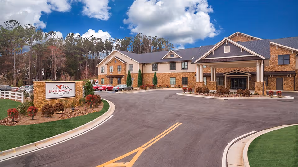Exterior view of Brickmont at Acworth assisted living facility showing a two-story building with a covered entrance, a curved driveway, landscaped greenery, and a sign with the facility name near the road.