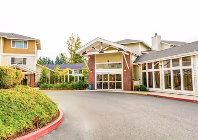 Exterior view of the Courtyard Fountains senior living facility entrance with a driveway, landscaped greenery, and multiple connected buildings featuring large windows and a covered entryway.