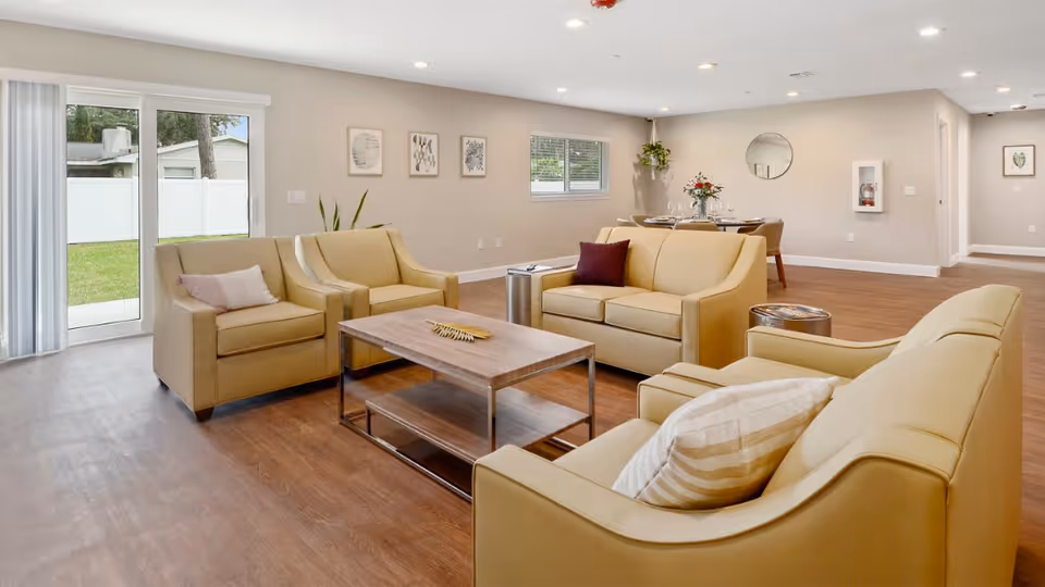 A bright and spacious living room area with beige leather sofas and armchairs arranged around a wooden coffee table. The room has wood flooring, a sliding glass door leading to a green outdoor space, and a dining table with chairs in the background. The walls are decorated with framed artwork and a round mirror.