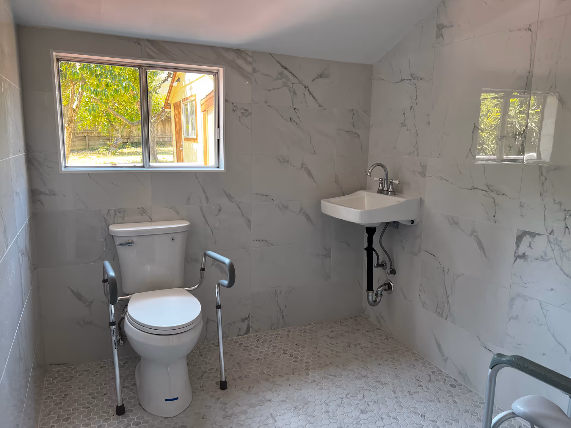A bathroom with white marble-patterned tiled walls and hexagonal tiled floor. There is a white toilet with safety rails on both sides positioned under a window showing an outdoor view with trees and a wooden fence. A small white wall-mounted sink with a silver faucet is on the right side.