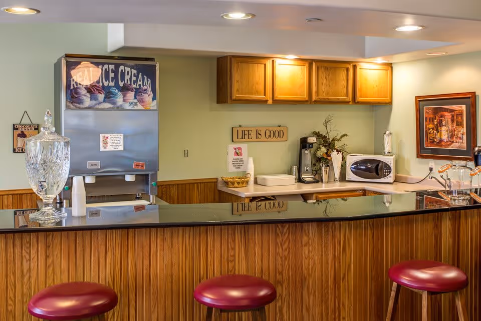 Interior view of a small kitchen or snack bar area with a wooden counter and three red cushioned stools. On the counter, there is a large glass beverage dispenser. Behind the counter, there is an ice cream machine with a sign that says 'REAL ICE CREAM', wooden cabinets, a coffee maker, a microwave, and a decorative sign on the wall that reads 'LIFE IS GOOD'. A framed picture hangs on the wall to the right.