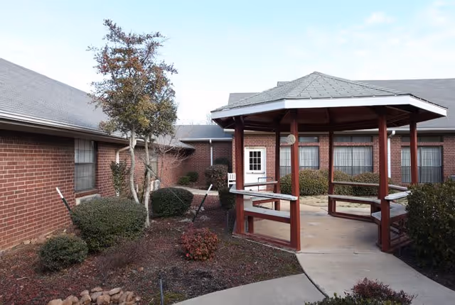 Outdoor courtyard area at Lamar Place featuring a red wooden gazebo with benches, surrounded by brick buildings, bushes, small trees, and a paved walkway under a clear sky.