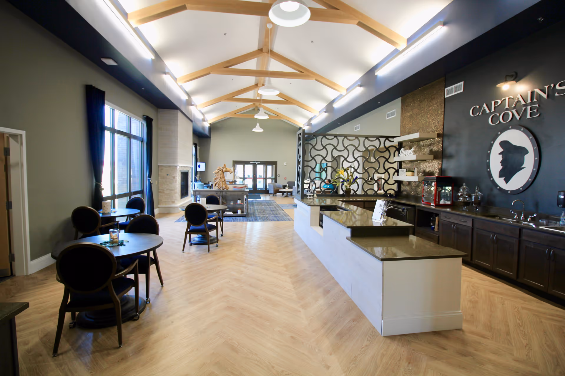 Spacious senior living common area with a high beamed ceiling, round dining tables, and a long serving counter under a 'Captain's Cove' sign.