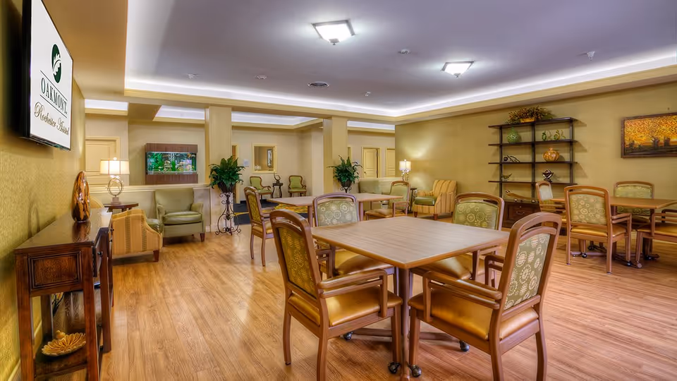 A spacious and well-lit common area in an assisted living facility featuring multiple wooden tables with cushioned chairs arranged around them. The room has warm yellow walls, wood flooring, decorative plants, a fish tank, and shelves with decorative items. There are also several armchairs and lamps providing a cozy atmosphere.