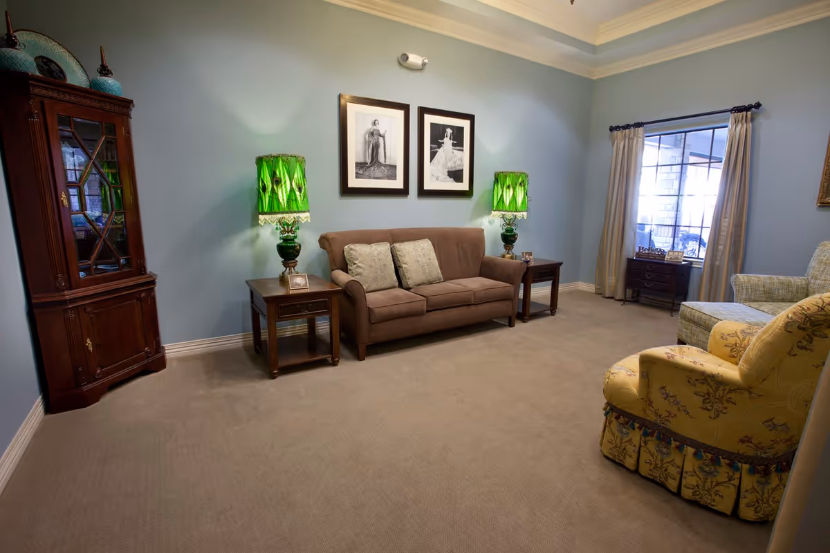 A well-appointed living room with a central sofa between two side tables topped by green lamps, framed wall art, upholstered chairs, and a display cabinet.