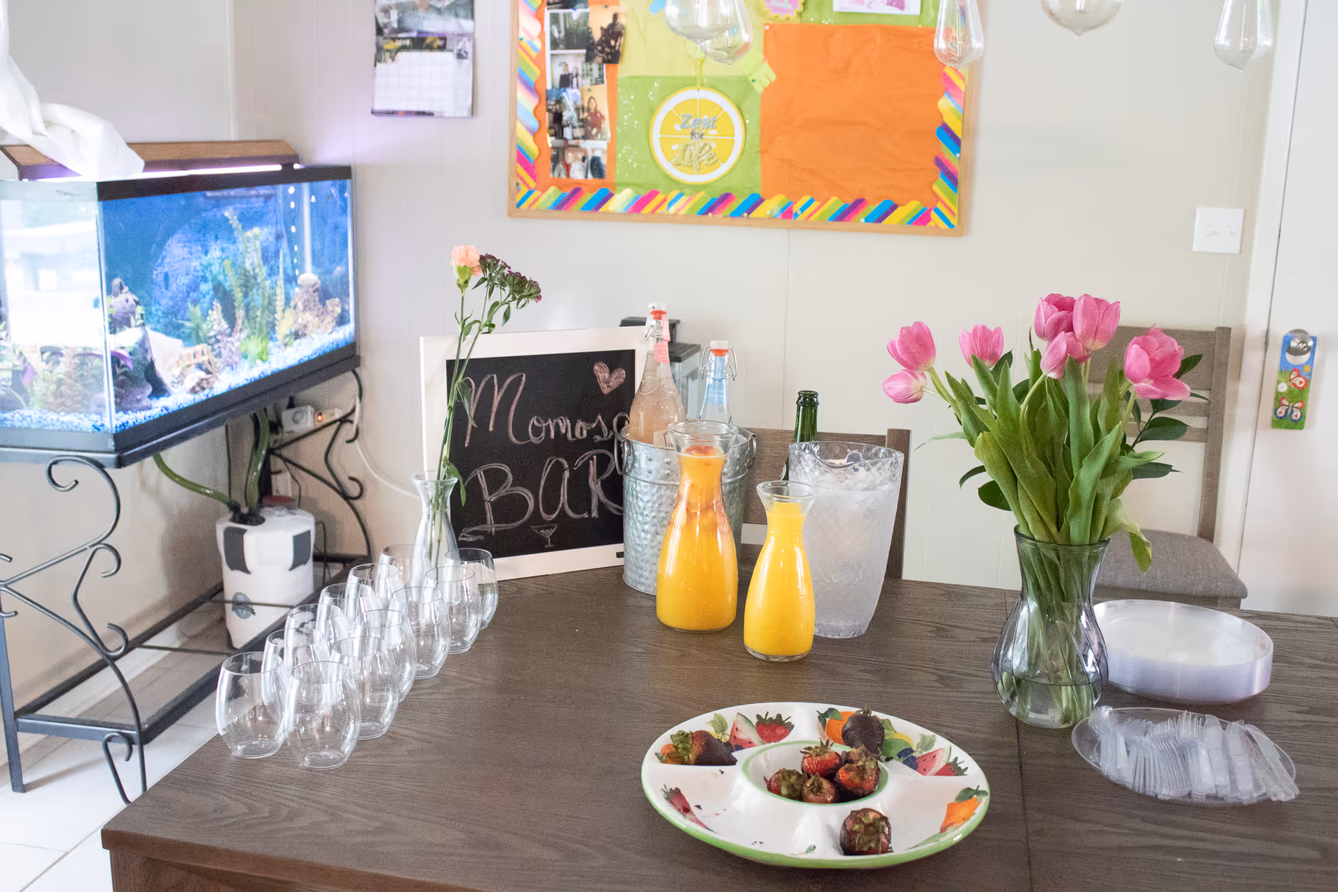 Dining table set with stemless glasses, carafes of orange juice, a vase of pink tulips and a small snack plate with a fish tank in the background.