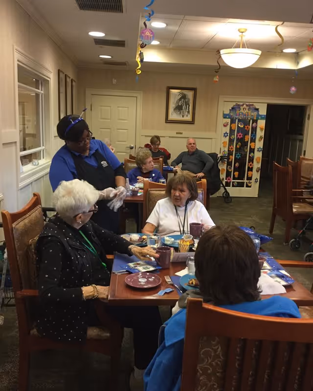 A group of elderly people seated around tables in a dining area at a senior living facility. A staff member wearing a blue uniform and gloves is interacting with one of the seated women. The room is decorated with hanging streamers and a door in the background is adorned with colorful decorations.