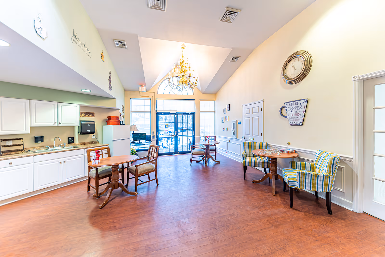 A bright and spacious common area in an assisted living facility featuring a kitchenette with white cabinets and a sink on the left, several small round tables with chairs and armchairs arranged around the room, a large chandelier hanging from a vaulted ceiling, and large windows and glass doors letting in natural light at the far end. The walls are decorated with a clock, framed pictures, and a coffee-themed wall art.