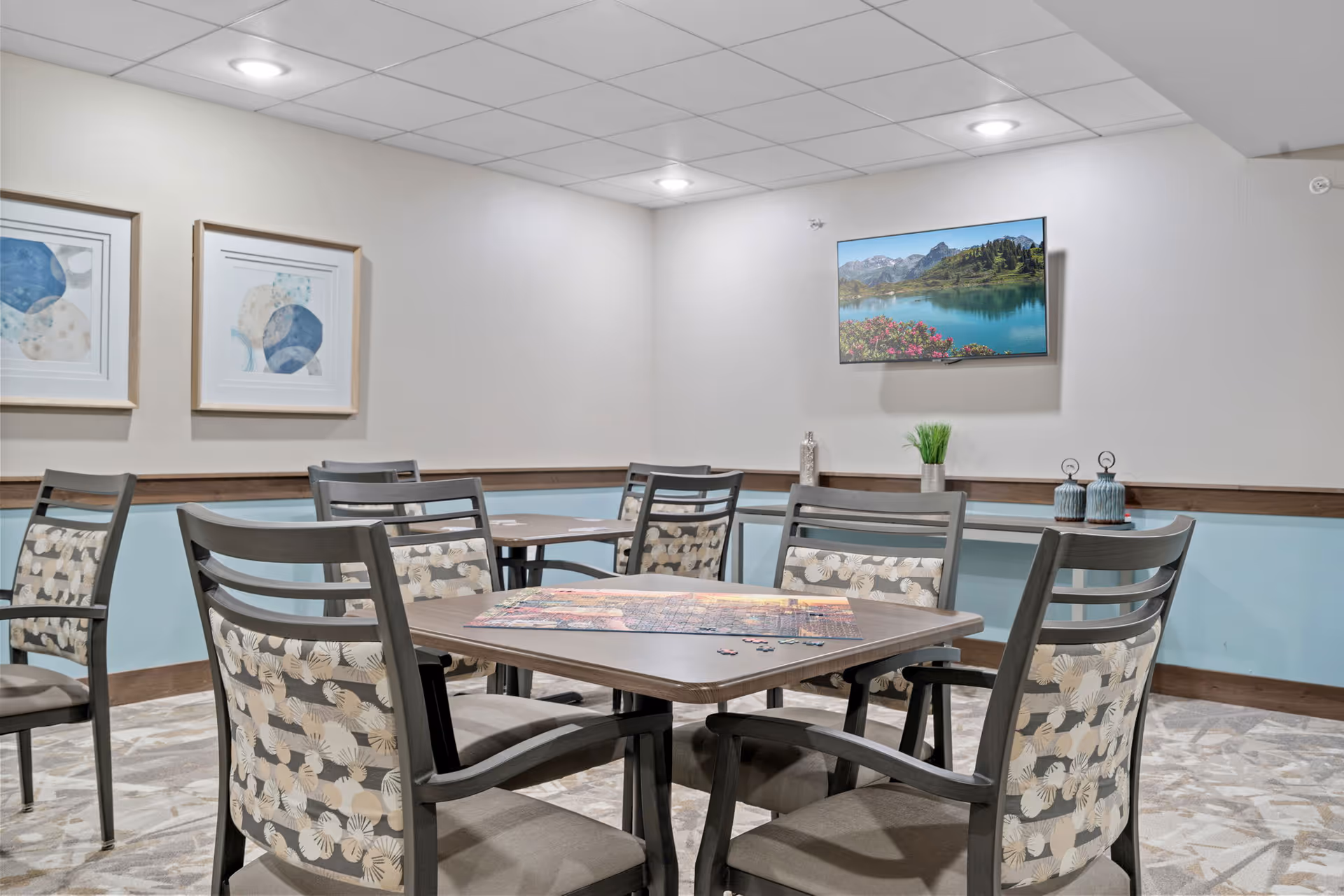 A bright senior living dining room with framed art and several tables and chairs.