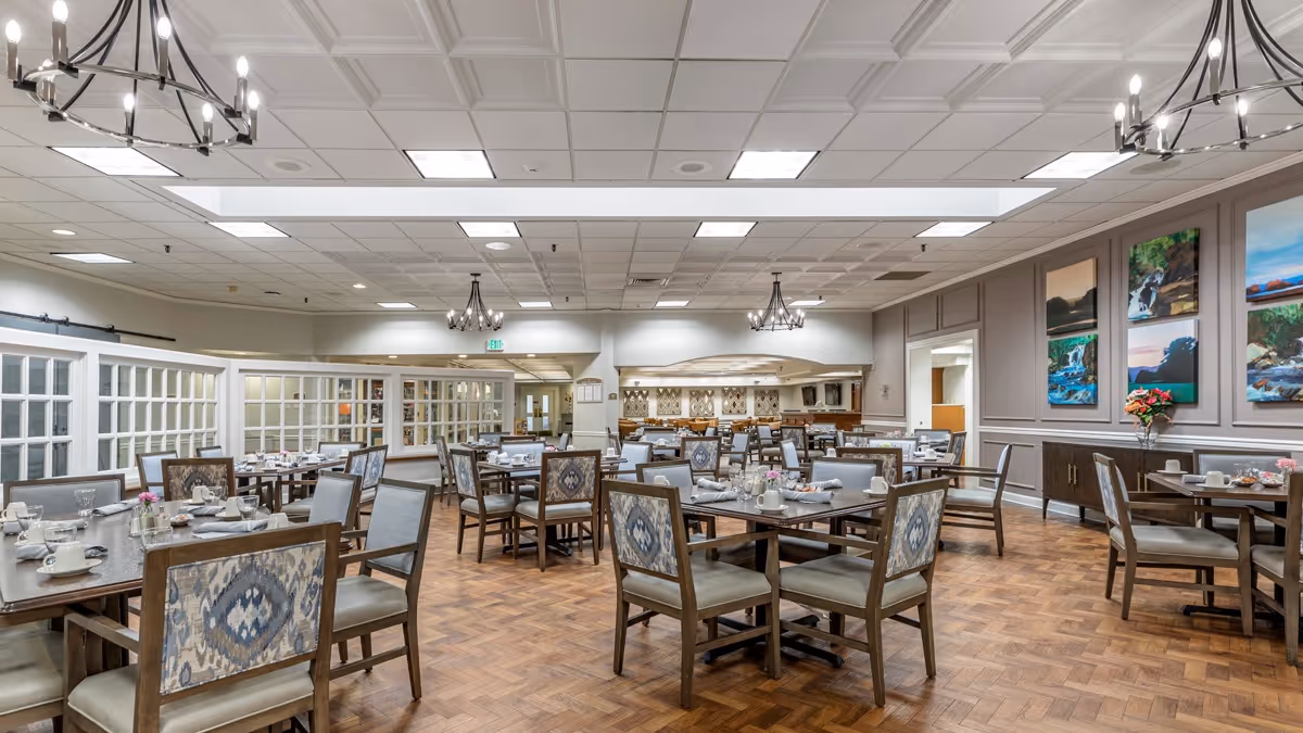 A spacious dining room with multiple tables and chairs arranged neatly. Each table is set with cups, plates, and napkins. The room features wooden parquet flooring, chandeliers hanging from the ceiling, and framed landscape artwork on the walls. There is a buffet area visible in the background.