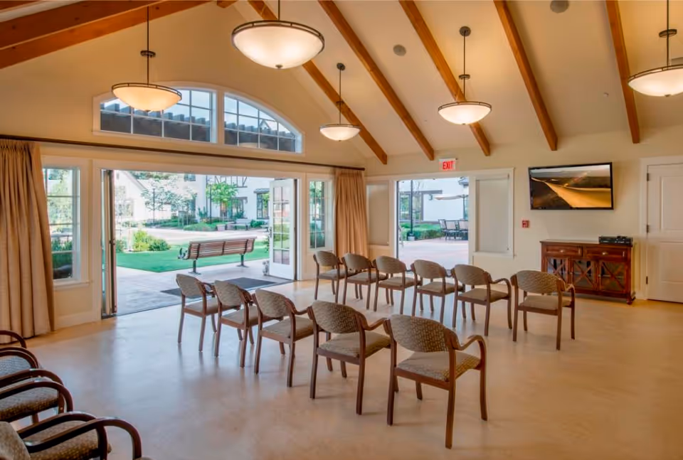Sunlit community room with rows of chairs facing a TV and wide open doors looking out to a courtyard and bench.
