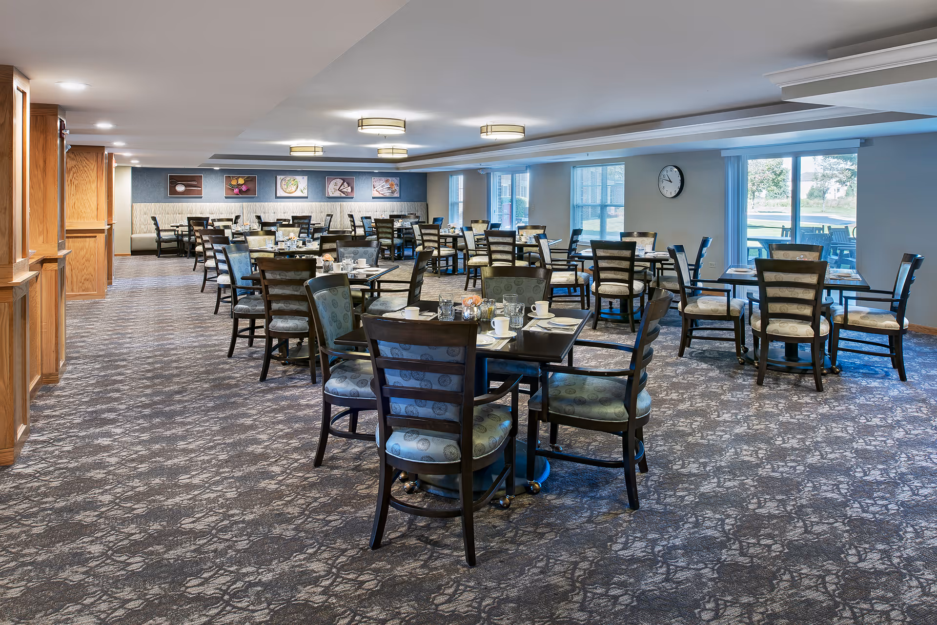 A spacious dining room with multiple tables and chairs arranged neatly. The tables are set with cups, glasses, and napkins. The room has large windows allowing natural light to enter, a wall clock, and framed pictures on the far wall. The carpet has a patterned design, and the ceiling features modern light fixtures.