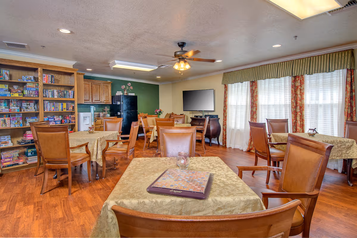 A cozy common room with several tables covered in beige tablecloths, surrounded by wooden chairs with leather cushions. A bookshelf filled with board games is visible on the left side, and a black refrigerator and wooden cabinets are in the background. The room has large windows with white curtains and red patterned drapes, a ceiling fan with lights, and a flat-screen TV mounted on the wall.