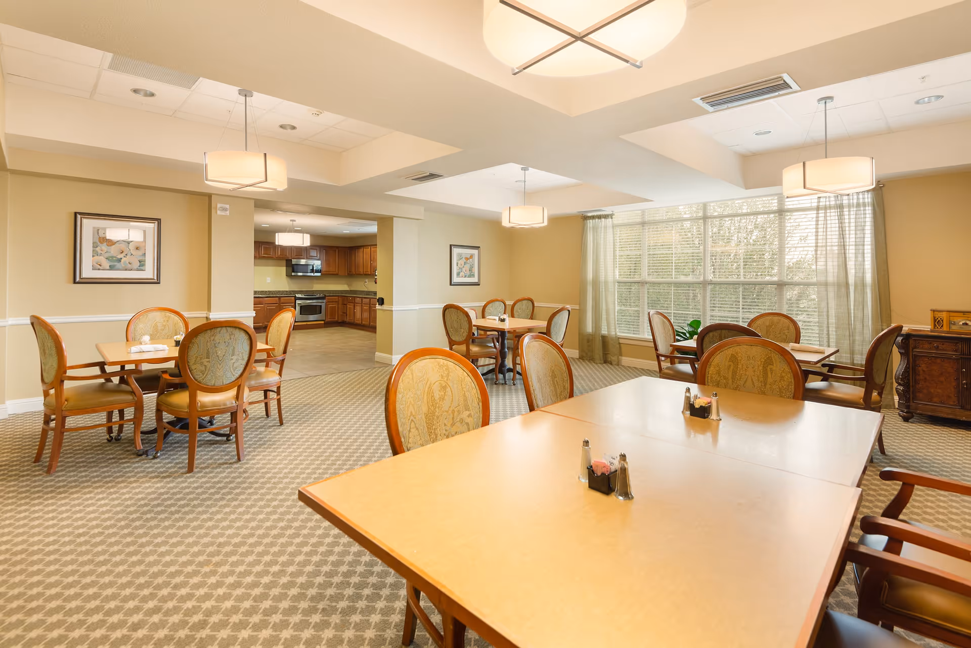 A bright dining room with several wooden tables and upholstered chairs arranged neatly. The room has large windows with sheer curtains allowing natural light to fill the space. There are pendant lights hanging from the ceiling and framed artwork on the walls. In the background, a kitchen area with wooden cabinets and stainless steel appliances is visible.