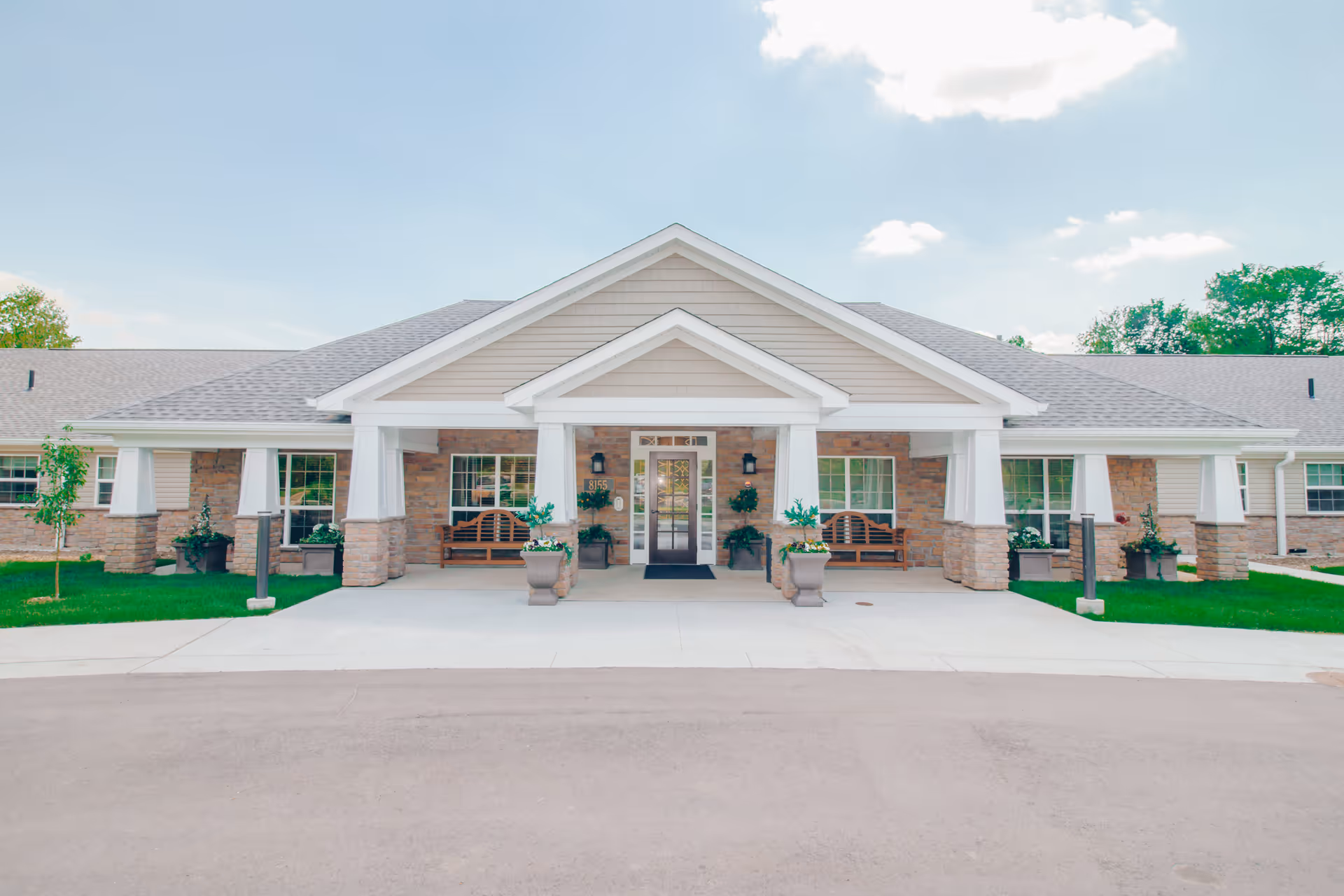 Front exterior view of a single-story senior living facility building with a covered entrance supported by white columns. The building has a combination of stone and siding on the facade, with benches and potted plants near the entrance. The sky is clear with a few clouds.