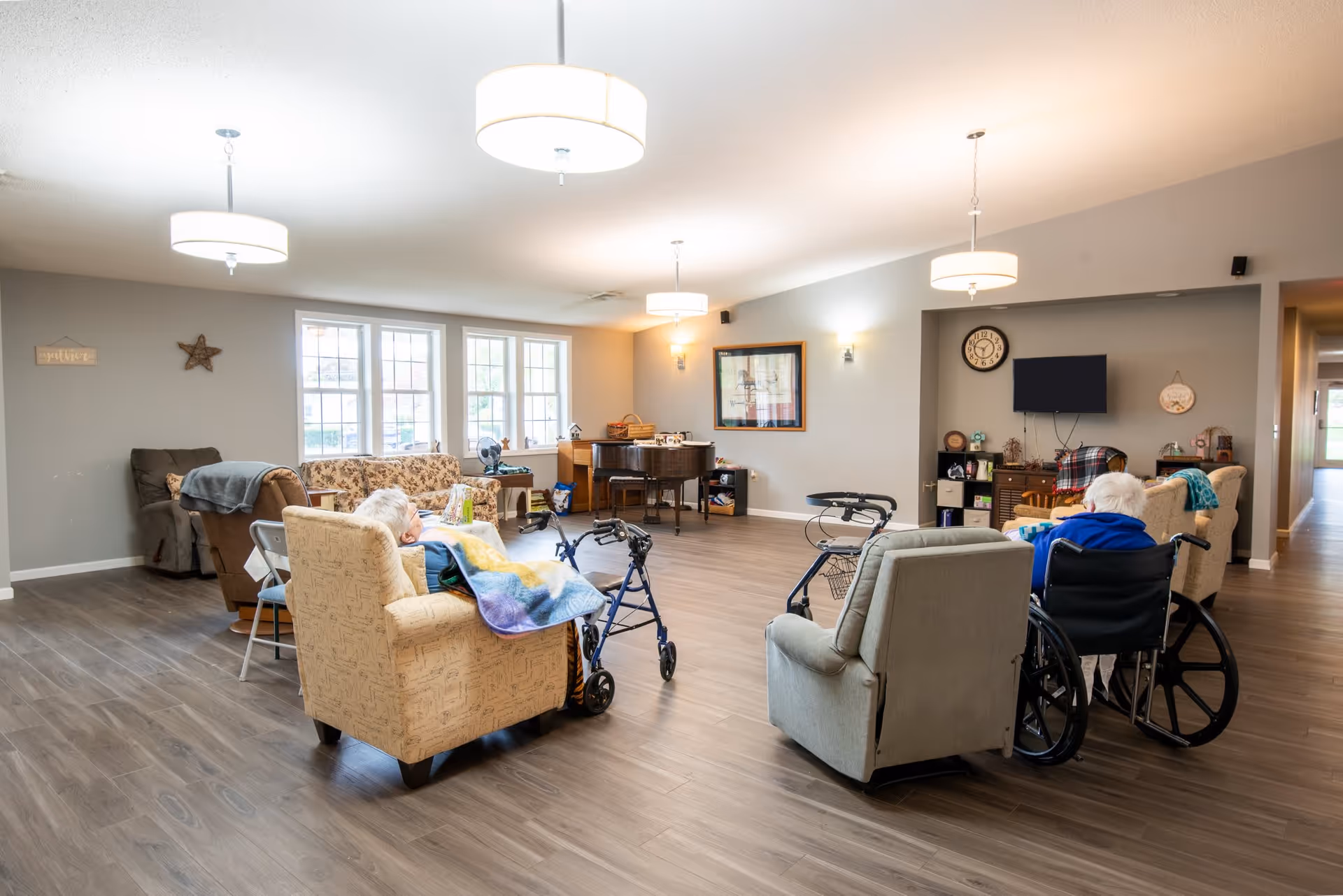 Spacious senior living common room with chairs, wheelchairs and walkers, residents seated, a piano and TV under pendant lights.