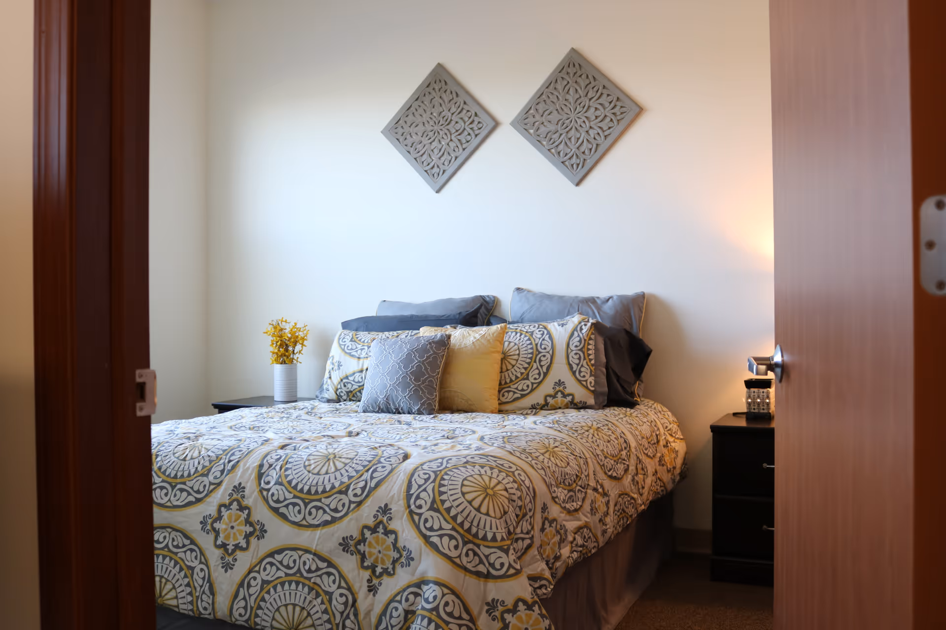 View into a neatly made bedroom with a patterned bedspread, decorative pillows, two wall plaques, and nightstands with a lamp and vase.