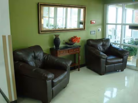 Two dark leather armchairs flank a small wooden table in a green senior living lounge.