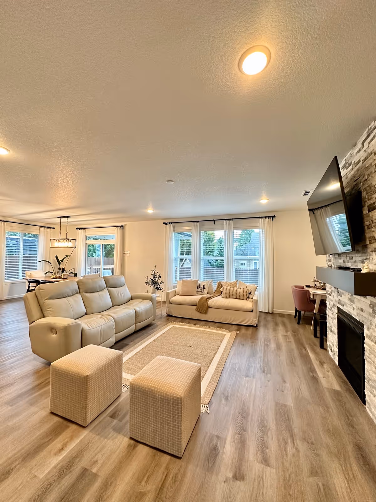 A spacious living room with light wood flooring, featuring a beige leather reclining sofa, a cream fabric sofa with decorative pillows, two small beige ottomans, a rectangular area rug, and a wall-mounted flat-screen TV above a modern stone fireplace. Large windows with white curtains allow natural light to fill the room, and a dining area with a table and chairs is visible in the background.