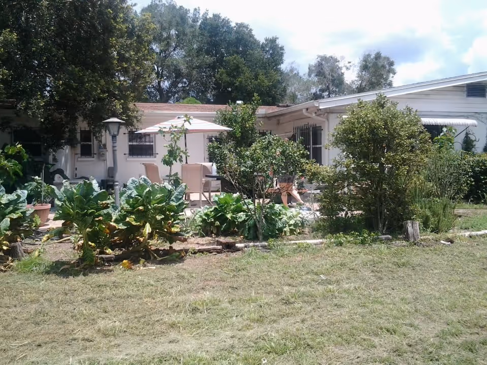Outdoor patio area at Sweet Water Grand The Bridges Assisted Living with a white building in the background, surrounded by green plants and trees. There is a table with chairs and an umbrella on the patio.