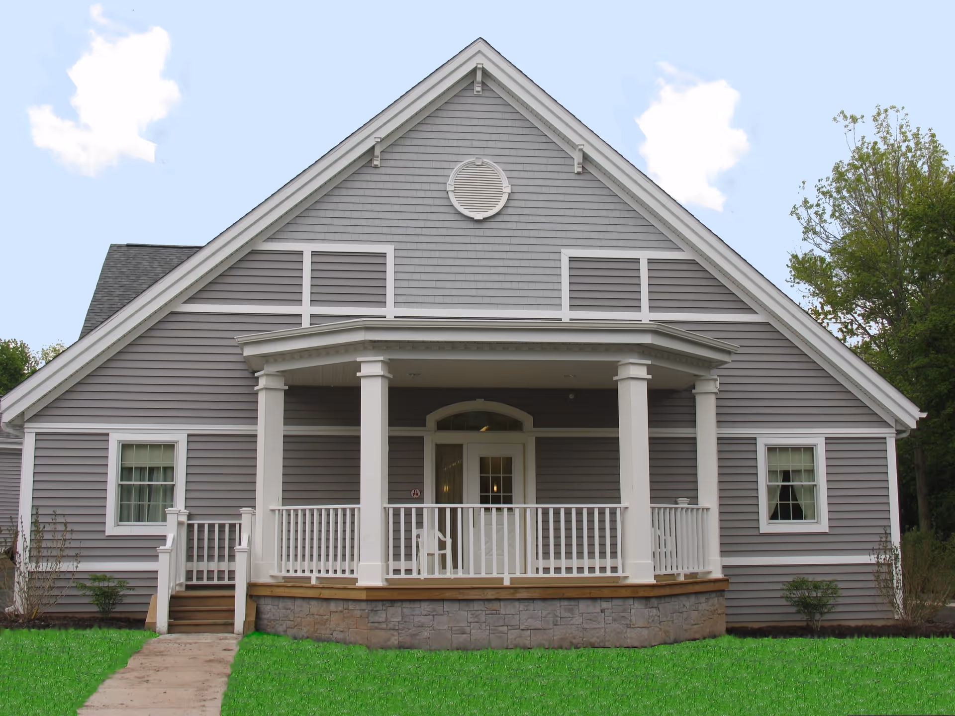 Front exterior view of a gray building with white trim and a small porch with white railings and columns. The building has a steep gabled roof and two windows on either side of the porch. There is a green lawn in front and trees in the background under a partly cloudy sky.