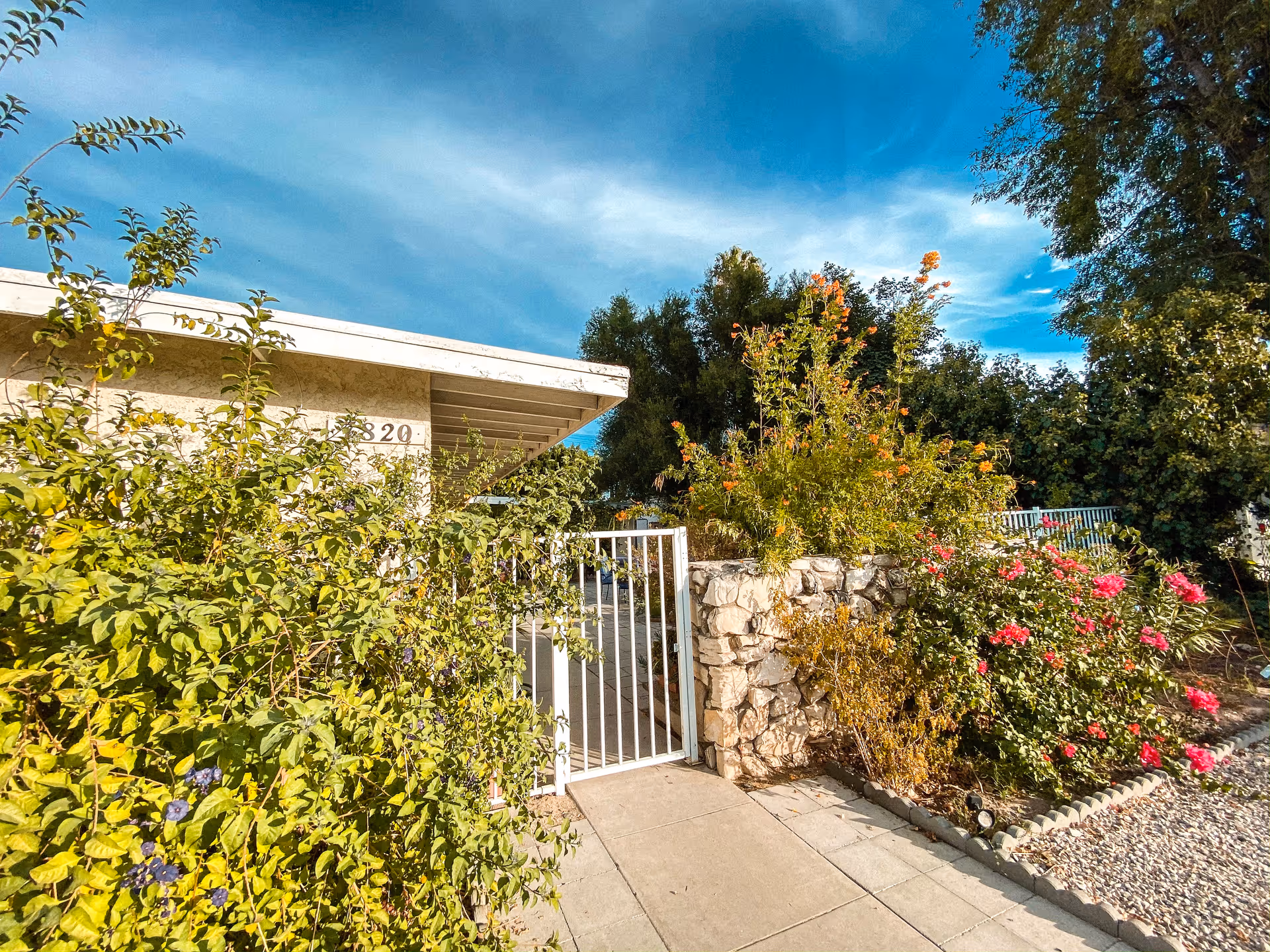 Gated entrance and stone planter with flowering shrubs in front of a single-story building under a blue sky.