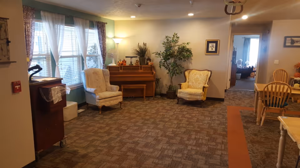 A cozy assisted living common area with two upholstered armchairs, a wooden piano with decorative items on top, a tall floor lamp, a large potted plant, and a window with sheer white curtains and patterned drapes. There is a wooden dining table with chairs to the right and a hallway leading to another room in the background.