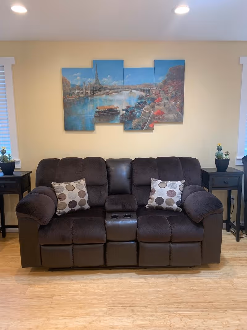 A cozy living room with a dark brown reclining sofa featuring two patterned cushions. On either side of the sofa are black side tables with small potted plants. Above the sofa is a four-panel wall art painting depicting a river scene with boats and a bridge under a blue sky. The room has light yellow walls and wooden flooring.
