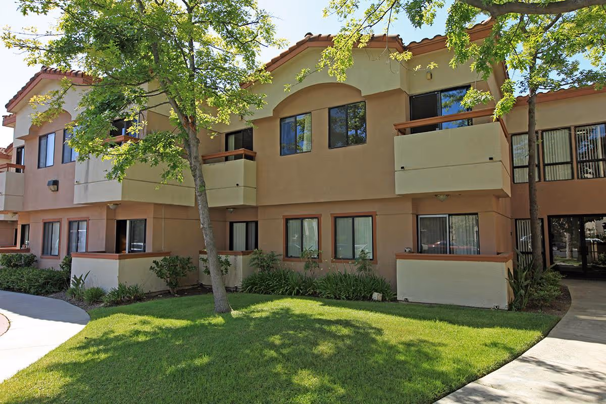 Two-story stucco apartment building with balconies facing a shaded grassy courtyard and walkway.